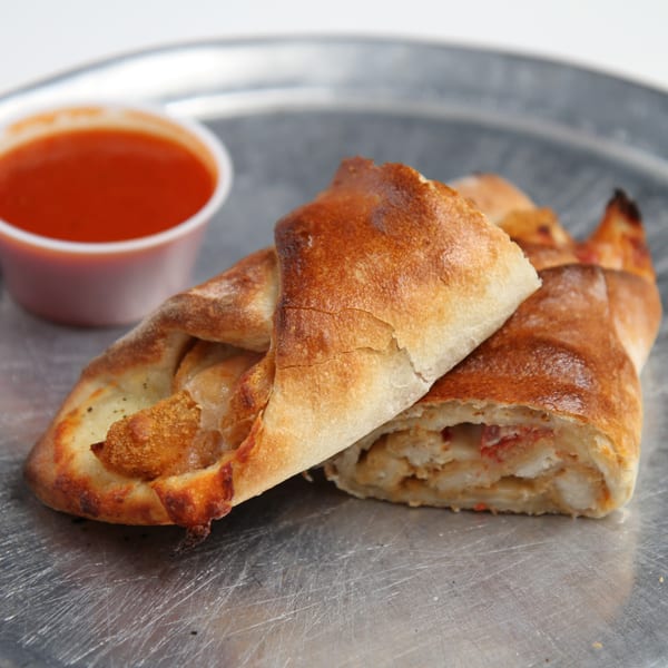 Two golden-brown strombolis with a side of red marinara sauce on a metal tray.