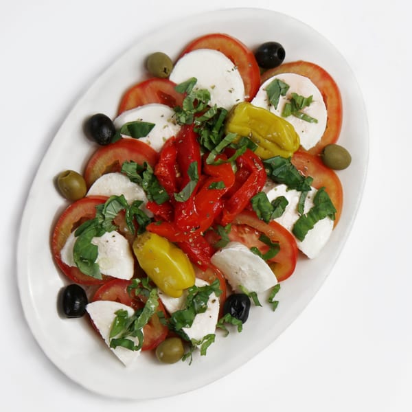 Caprese salad with sliced tomatoes, mozzarella, basil, olives, and peppers on a white platter.
