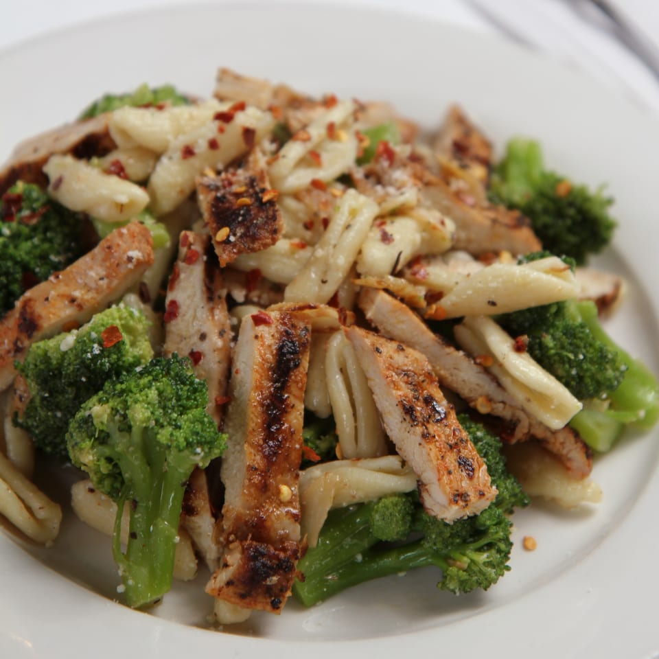 Grilled chicken and broccoli pasta with red pepper flakes on a white plate.