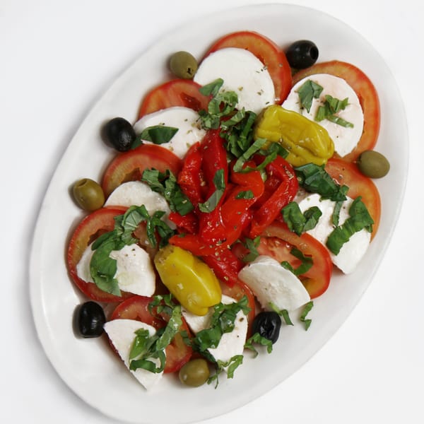 Caprese salad with sliced tomatoes, mozzarella, basil, olives, and roasted peppers on a white platter.